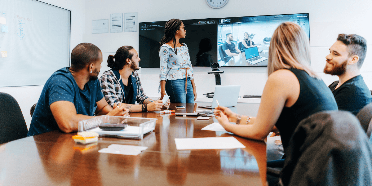 A woman with braided hair stands to present in a modern conference room, addressing four colleagues seated around a wooden table. Behind her, two wall-mounted screens display meeting content, including a video call with remote participants.